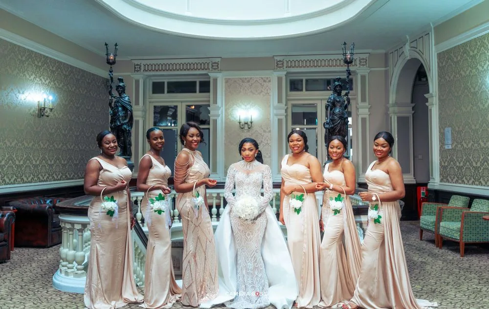 A bridal party poses in elegant champagne-colored gowns inside an ornate room with wall sconces and decorative molding.