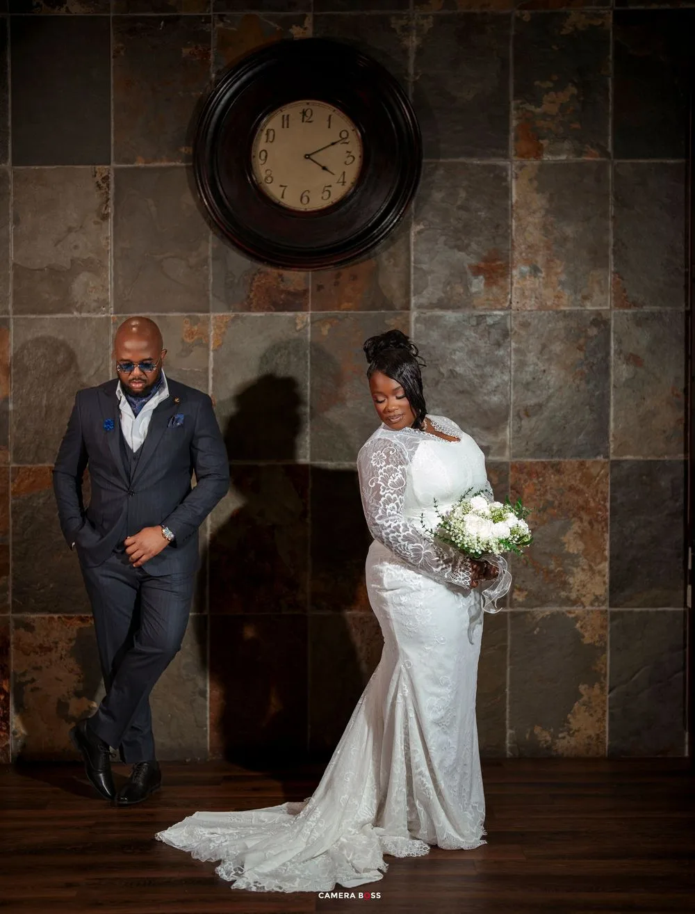 Elegant bride in lace gown holds white bouquet beside suited groom near rustic tile wall with vintage clock.