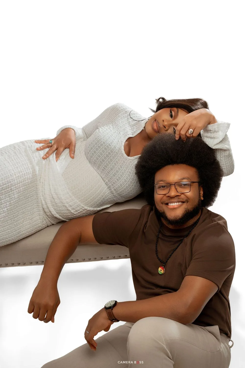 A pregnant woman in a white dress reclines on a bench while a smiling man in brown sits beside her.