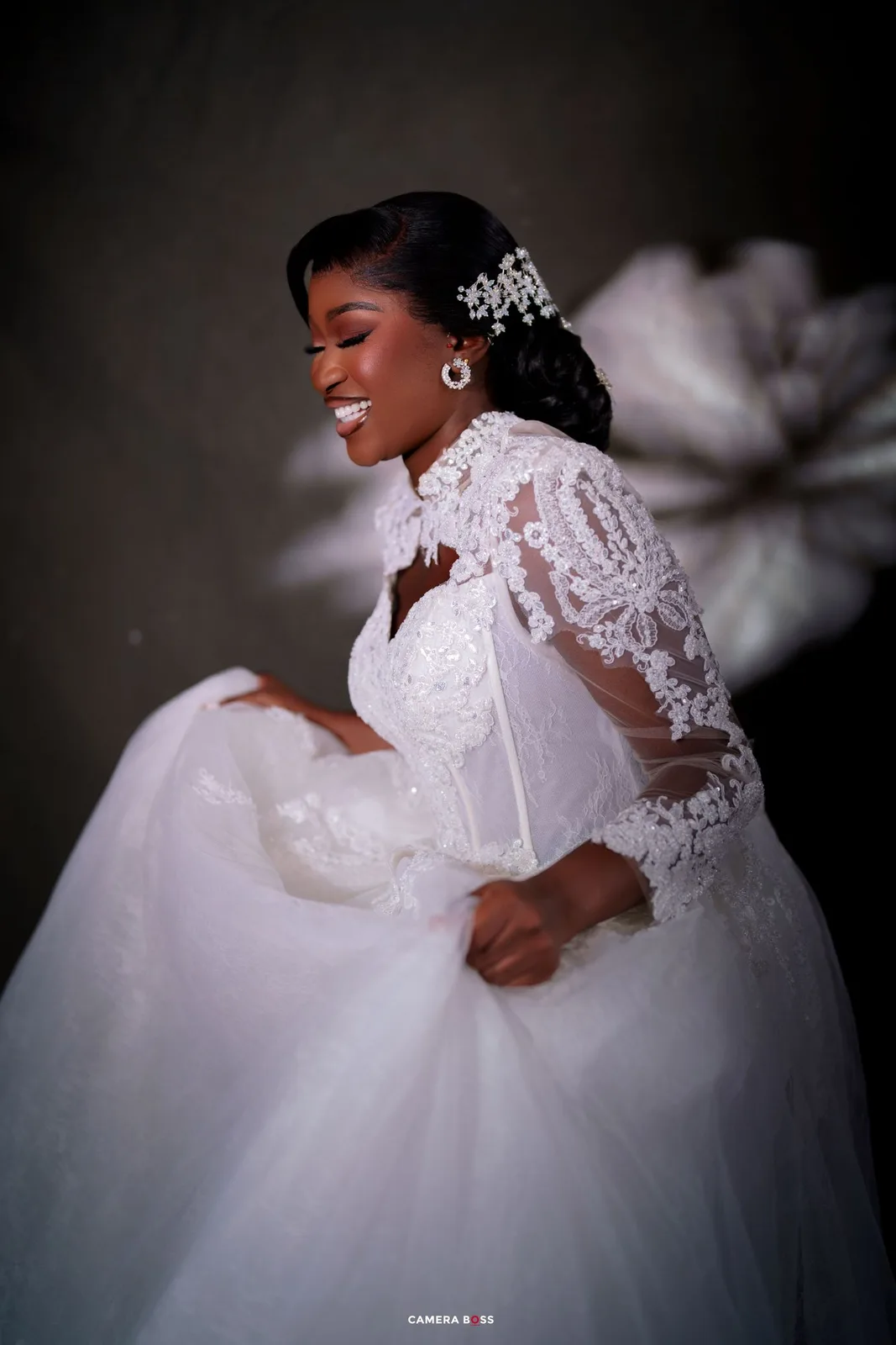 Smiling bride in white lace ball gown with sheer sleeves and crystal hair accessory poses joyfully.