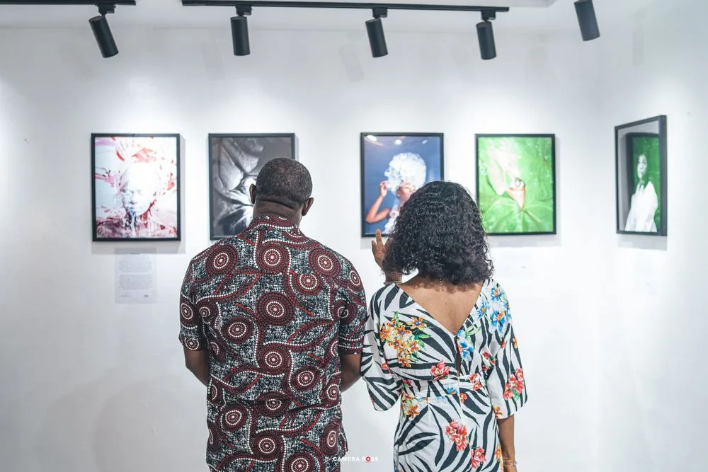 Two people wearing patterned clothing view colorful framed artwork displayed on a white gallery wall under track lighting.