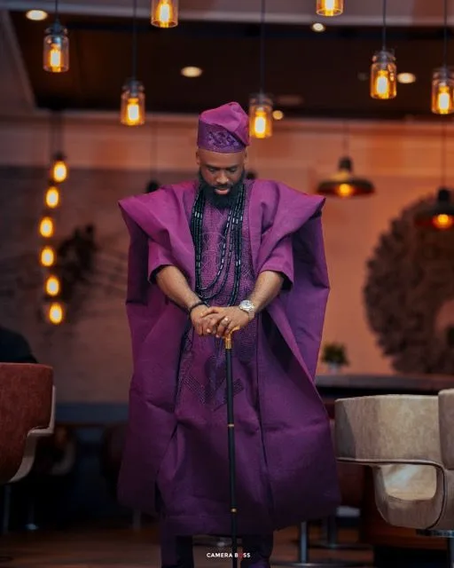A person wearing a striking purple traditional African agbada and cap stands elegantly in a warmly lit room with hanging lanterns.