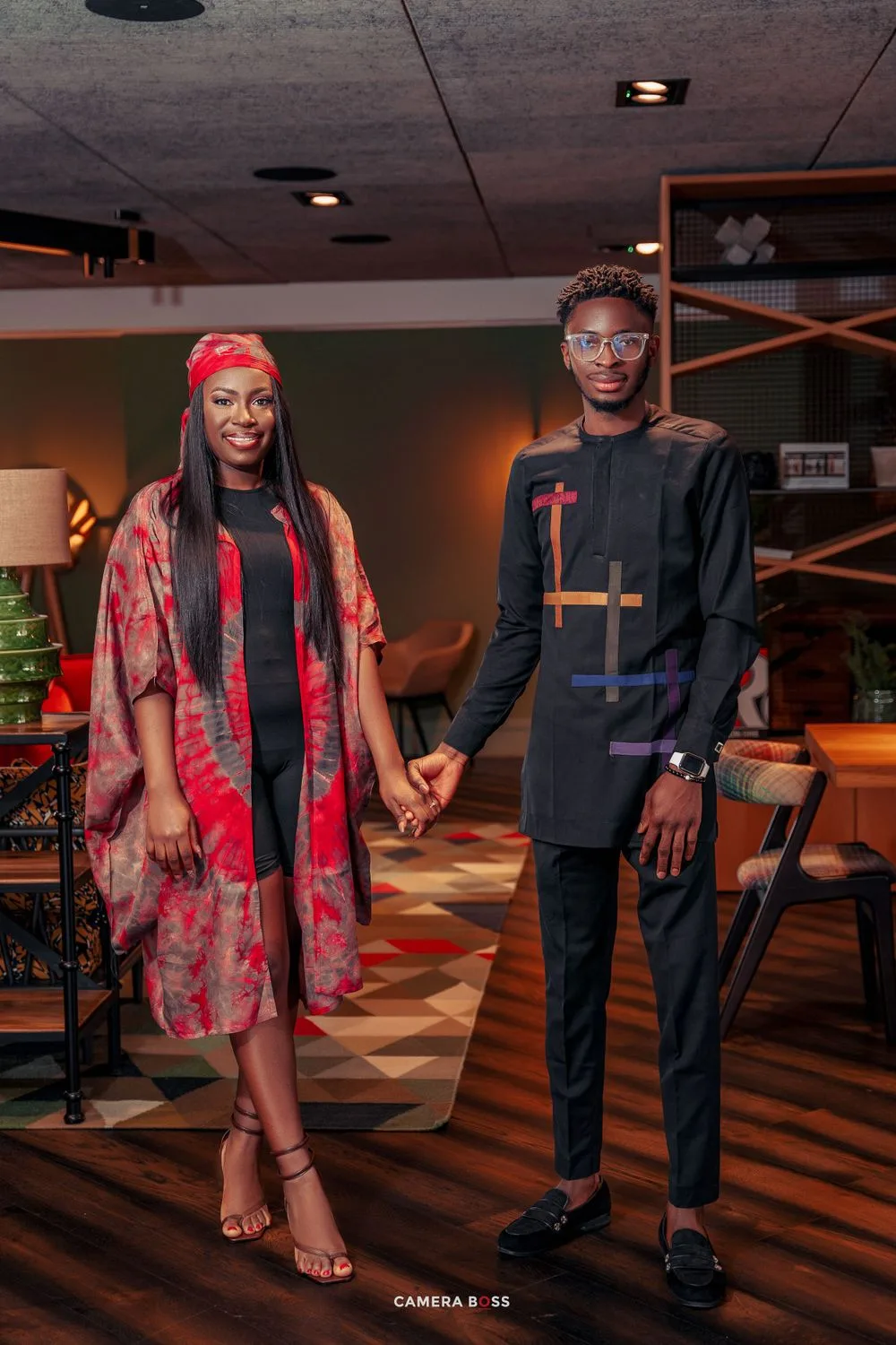 A couple in stylish formal wear poses together at an indoor venue with dark wood floors and ambient lighting.