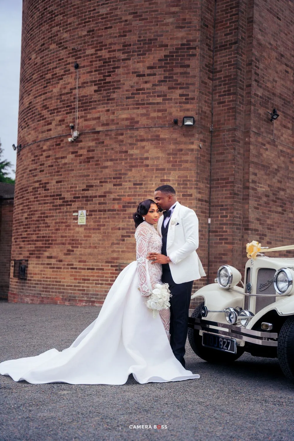 A couple shares a romantic moment in wedding attire next to a vintage car against a brick building backdrop.
