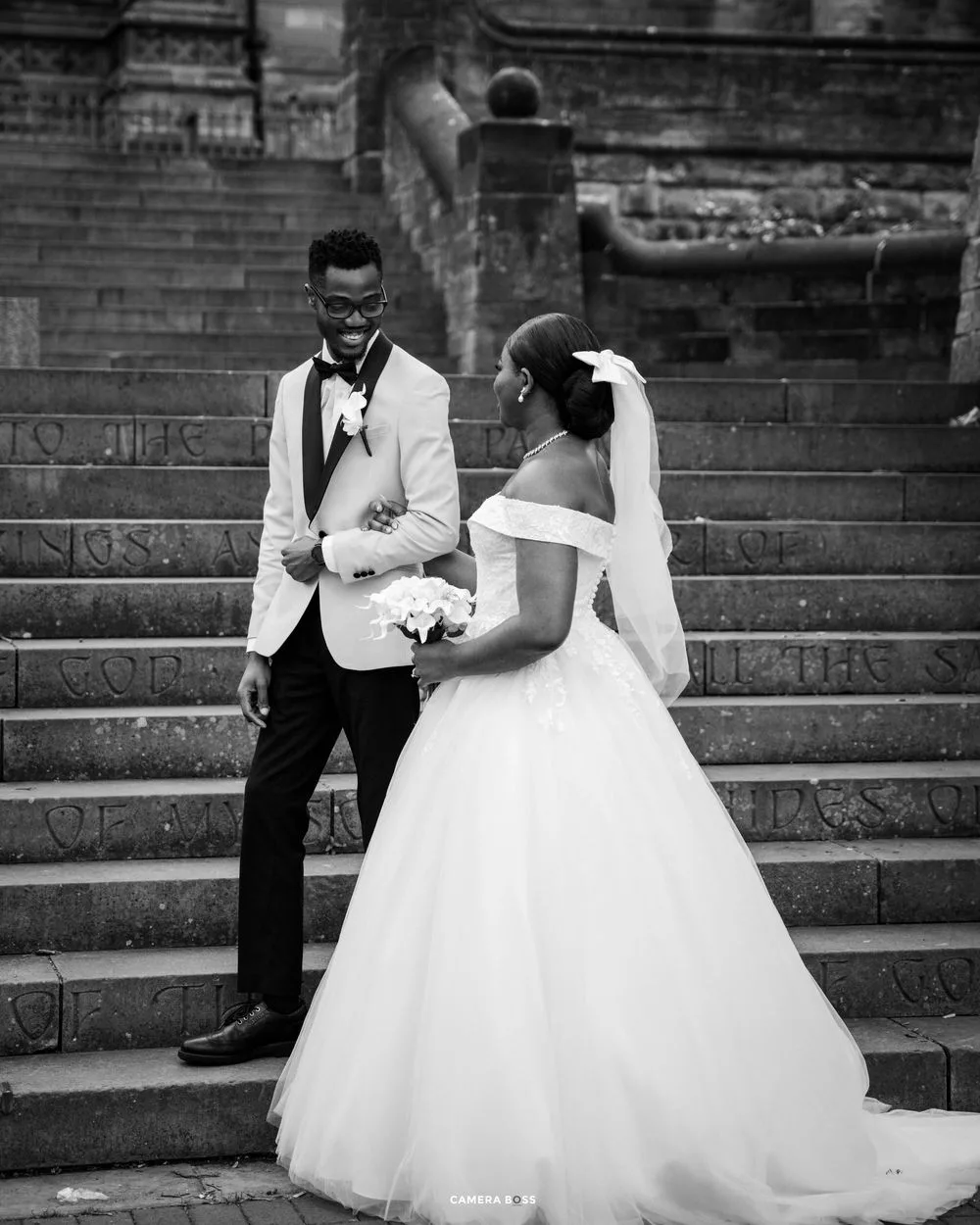 Black and white wedding photo of newlyweds sharing a joyful moment on stone stairs in formal attire.