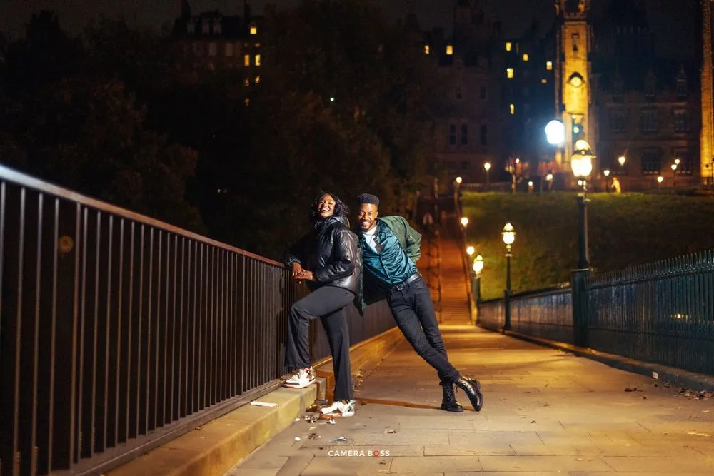 Two people pose playfully on a bridge railing at night with a city clock tower illuminated in the background.