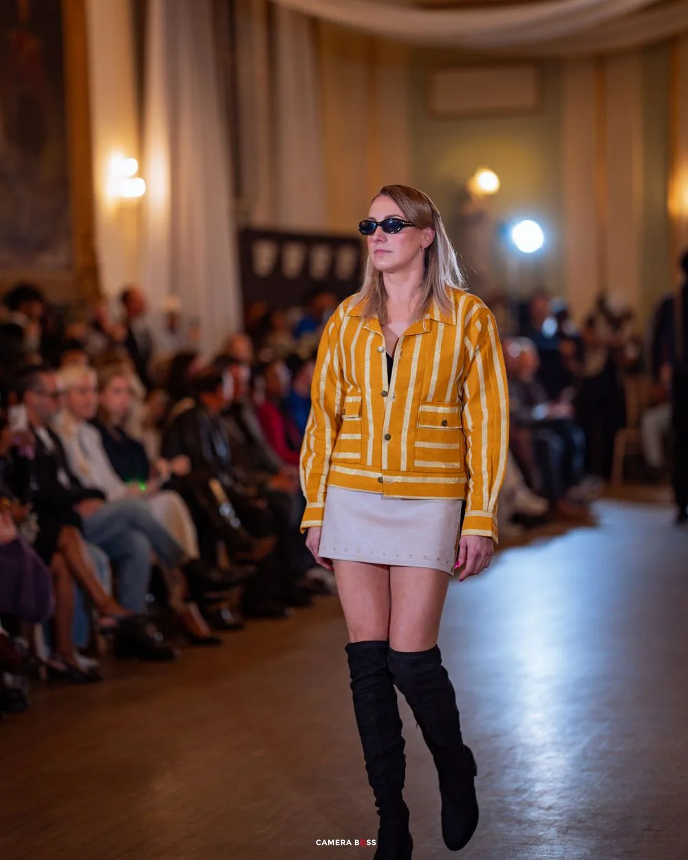 Model walks runway wearing yellow geometric patterned jacket, white skirt and black boots at indoor fashion show.
