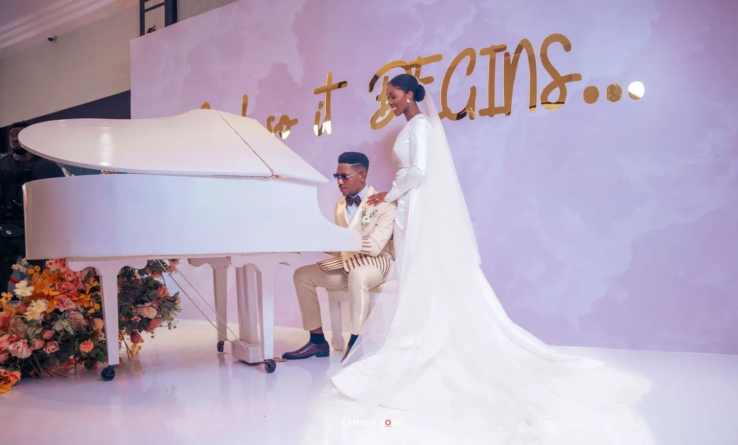 A romantic wedding scene with a white piano, floral decorations, and a flowing white bridal gown against a pink backdrop.