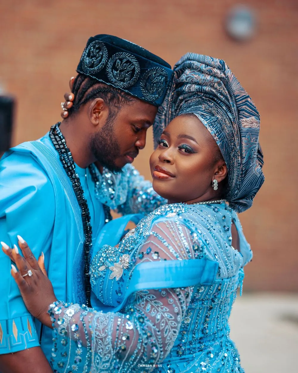 A couple in coordinated blue Nigerian wedding outfits share an intimate moment against a brick wall.