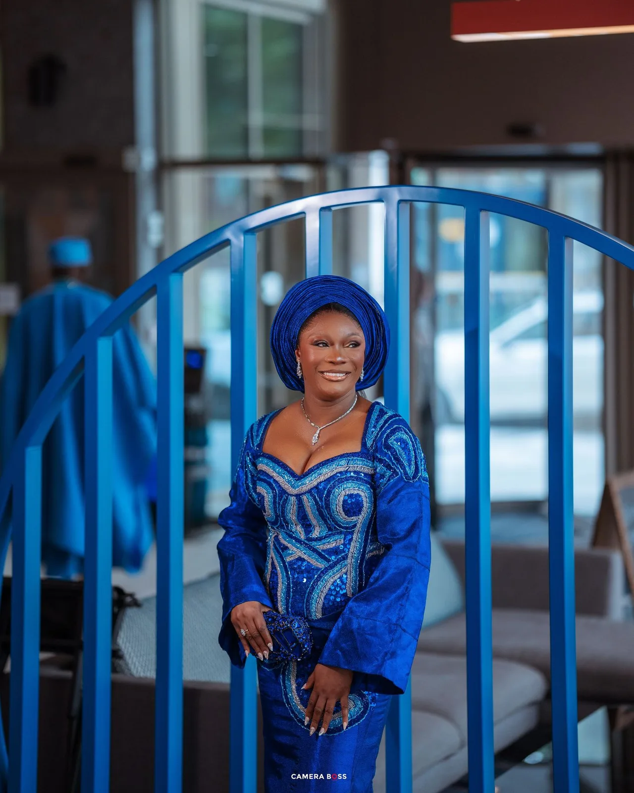 Individual wearing royal blue and white patterned traditional African dress poses by blue railing.