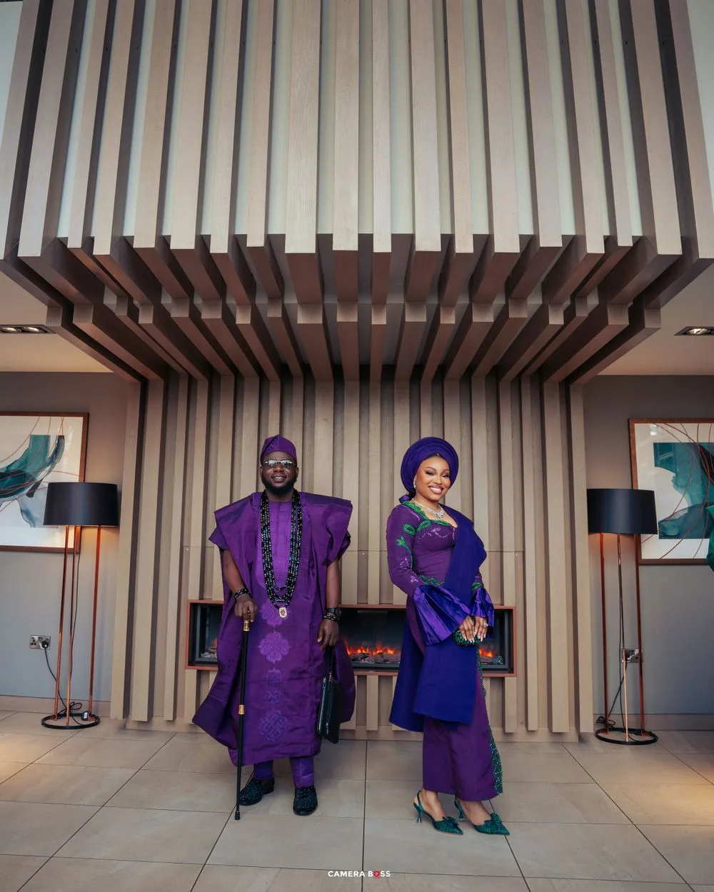 A couple in coordinating purple and blue traditional African attire poses elegantly in a modern interior setting.