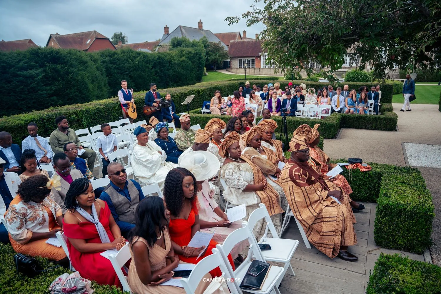 Outdoor wedding ceremony guests seated in white chairs along a manicured garden path with hedges and buildings in background.