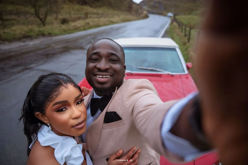 Couple taking a selfie in front of a red classic car on a scenic road, dressed in elegant formal attire.