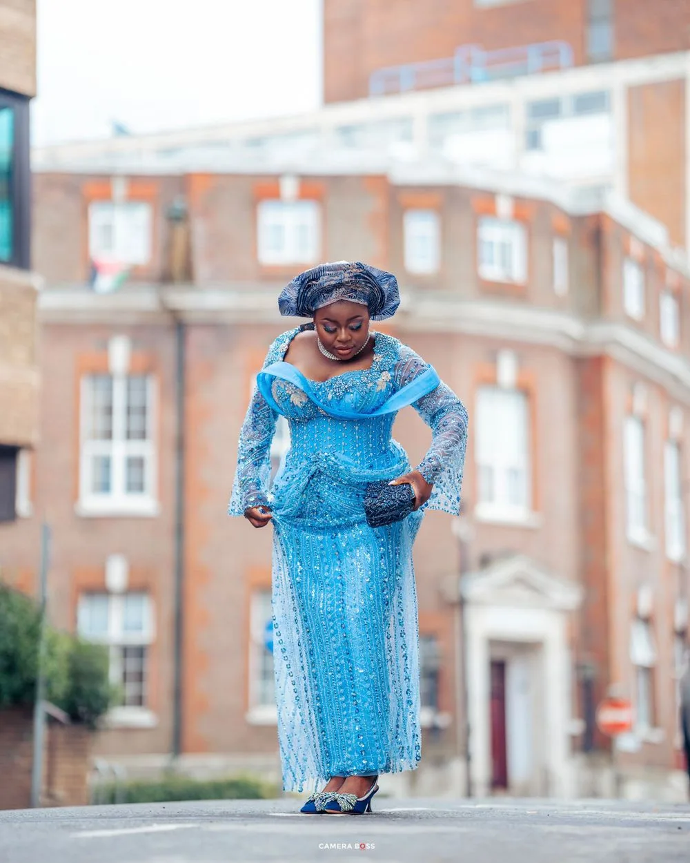 Woman in vibrant blue traditional African dress and headwrap poses gracefully on a city street with brick buildings behind her.