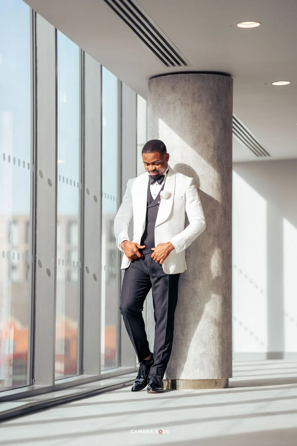 Well-dressed person in white blazer and dark pants poses near large windows in modern building with natural lighting.