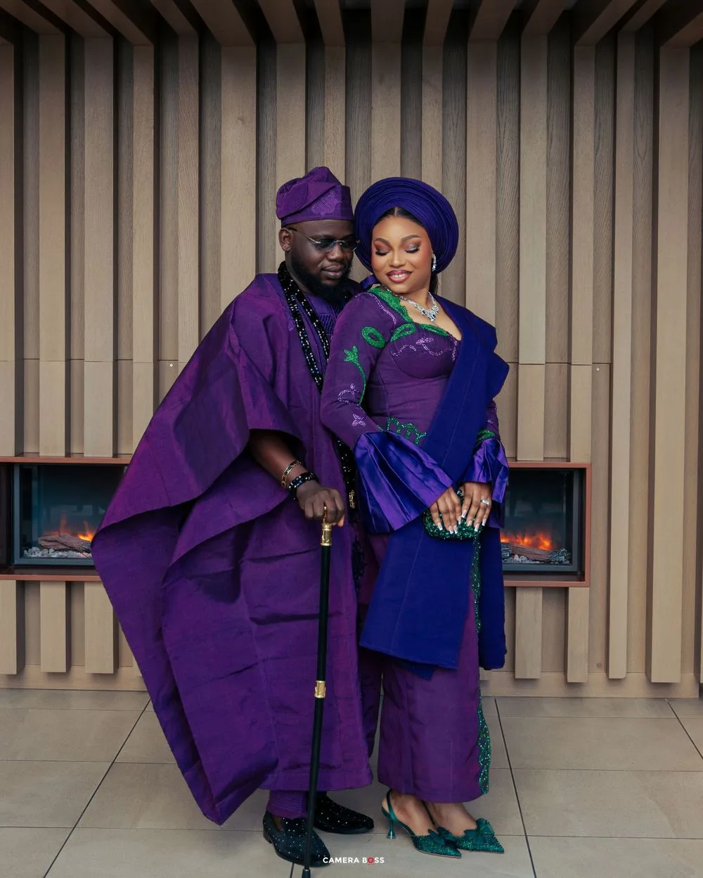 A couple in coordinating traditional African attire poses together, wearing royal purple and blue formal garments.
