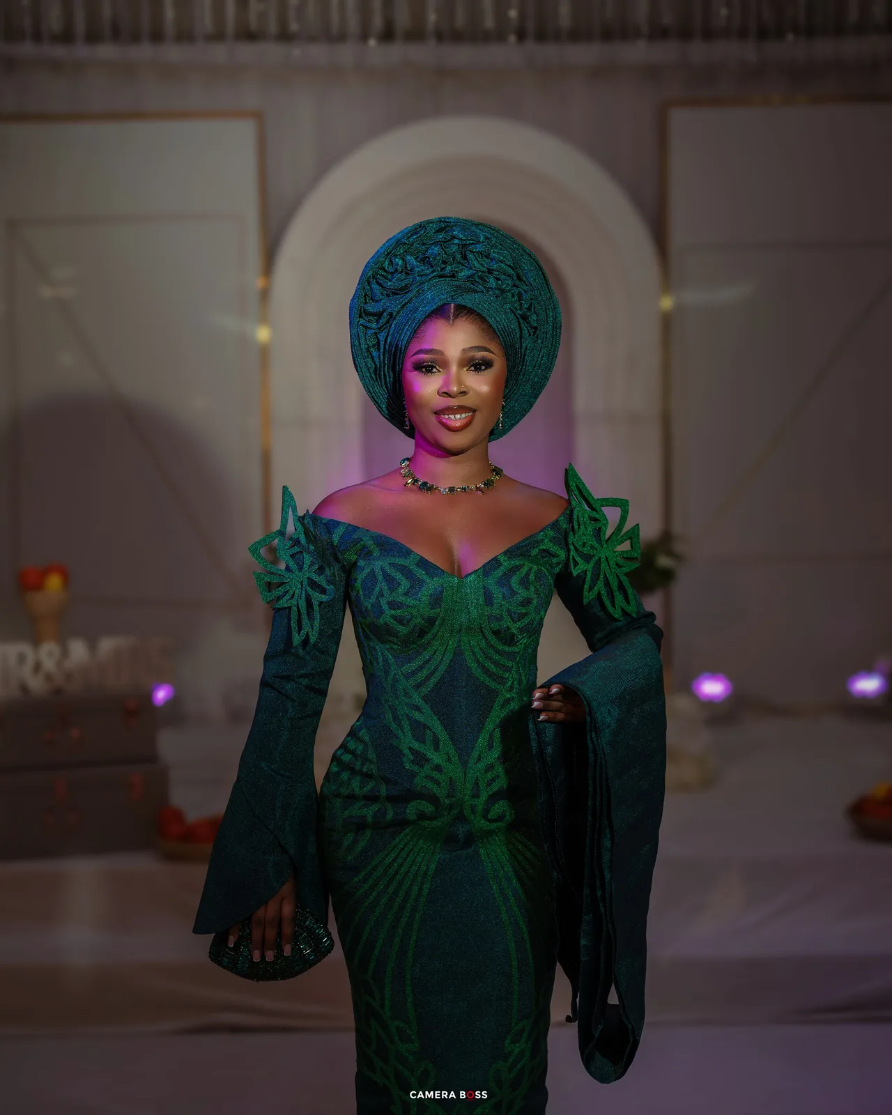 An elegant emerald green evening gown with an ornate headpiece and jeweled neckline glows under purple stage lighting.
