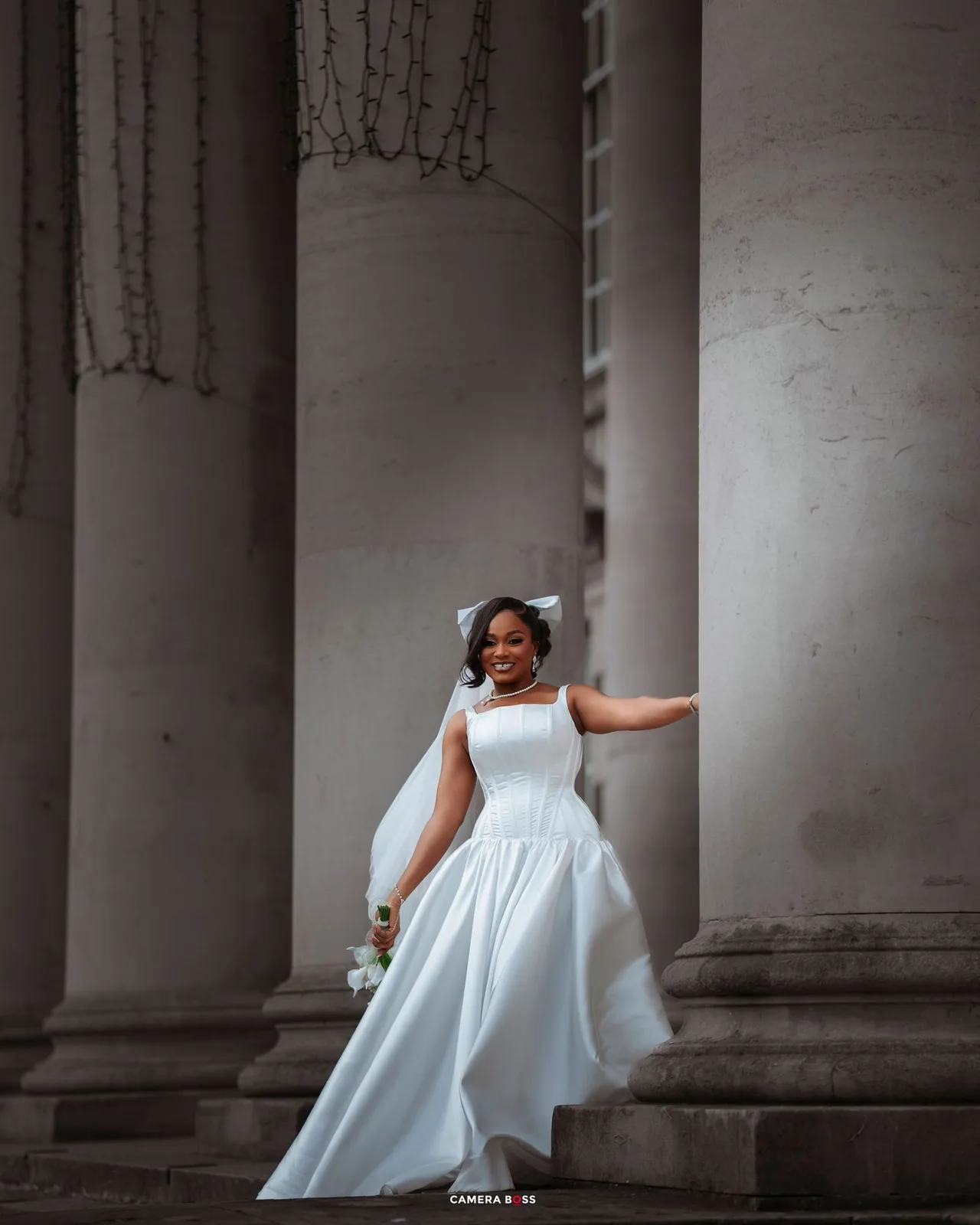 Bride in elegant white gown posing gracefully between grand stone columns, holding a bouquet.