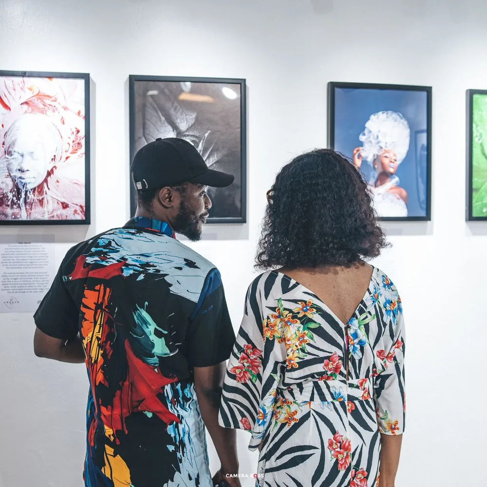 Two people wearing colorful patterned clothing view artwork displayed on a white gallery wall together.