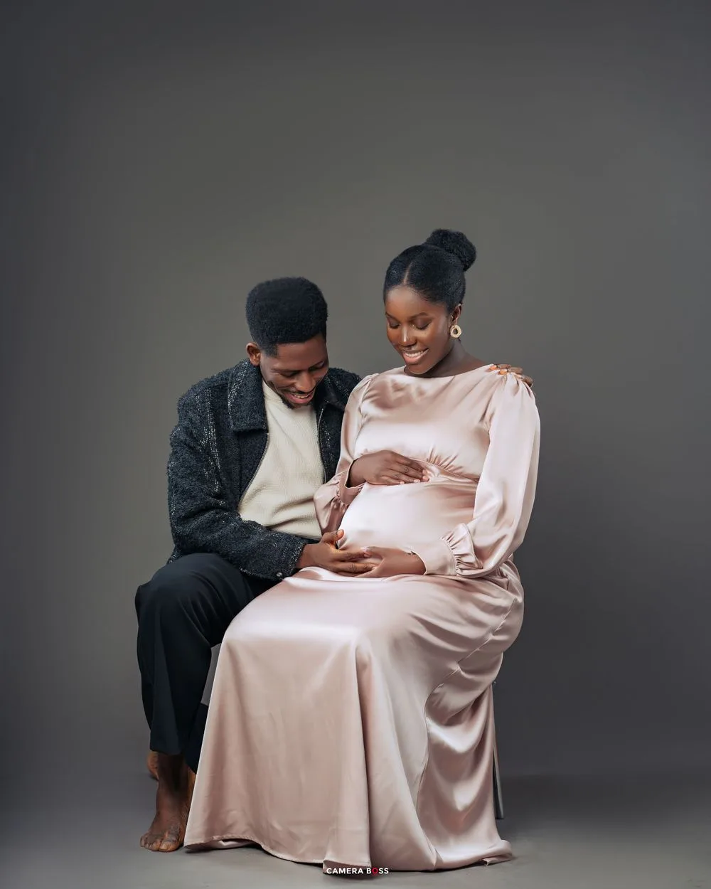 Intimate maternity photo featuring tender moment between expecting couple in light pink dress and dark sweater against gray backdrop.