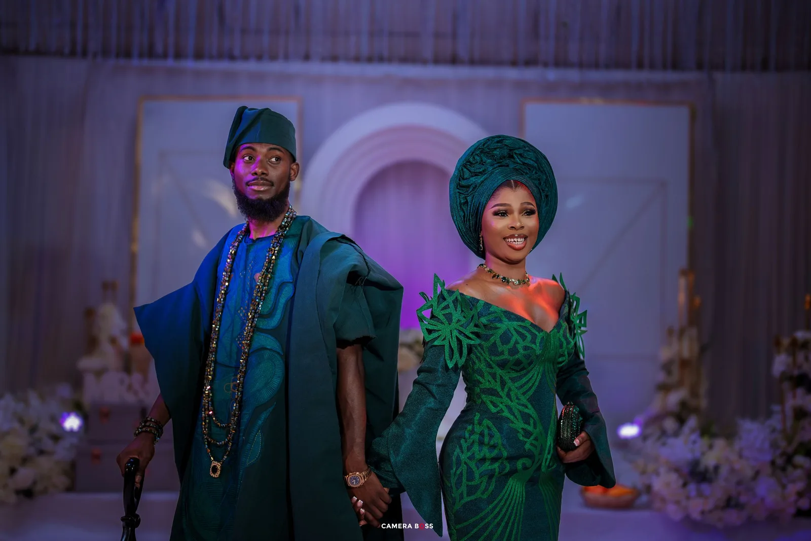 Two people in formal attire at an event with an arched backdrop, one wearing a green sequined gown and the other in blue.
