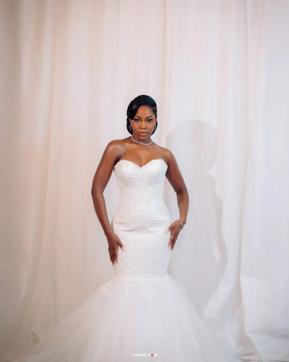 A bride poses in a stunning white strapless mermaid wedding gown with a tulle skirt against a light backdrop.
