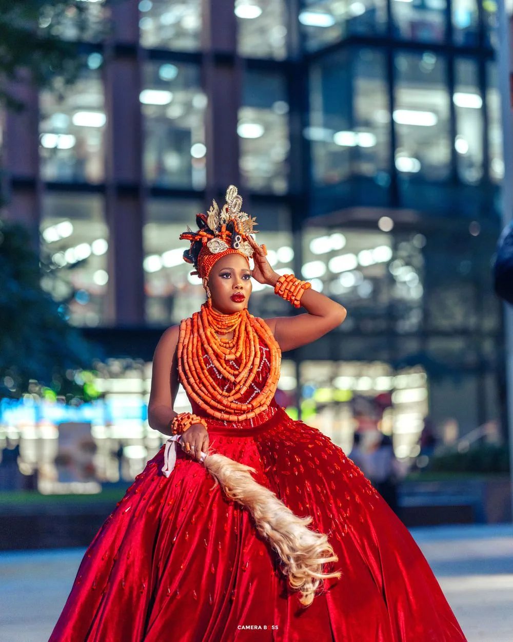 A dancer in a dramatic red ball gown and orange beaded necklaces poses elegantly against a modern glass building backdrop.