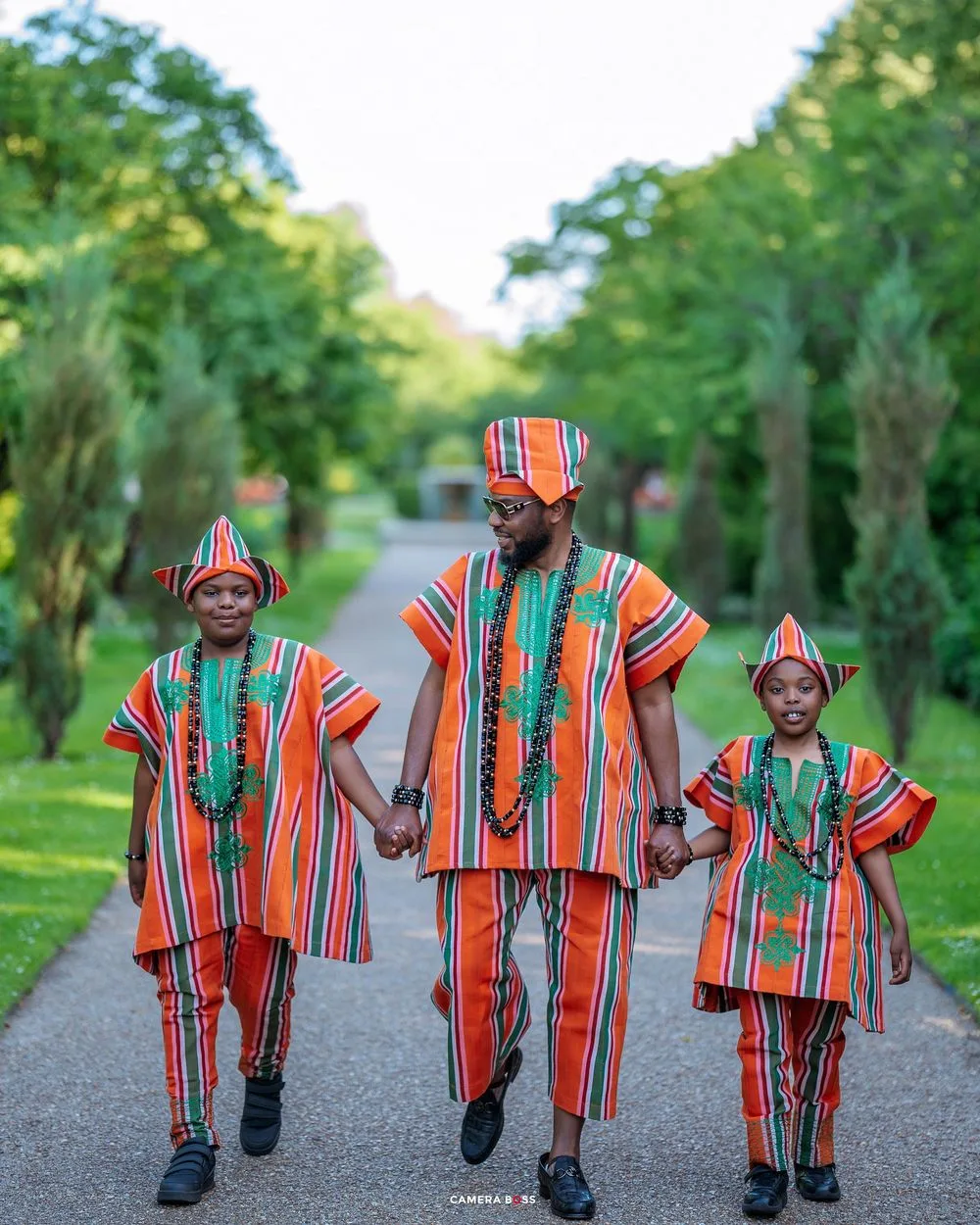 Three people walk hand in hand wearing vibrant orange and green striped traditional African outfits on a tree-lined path.