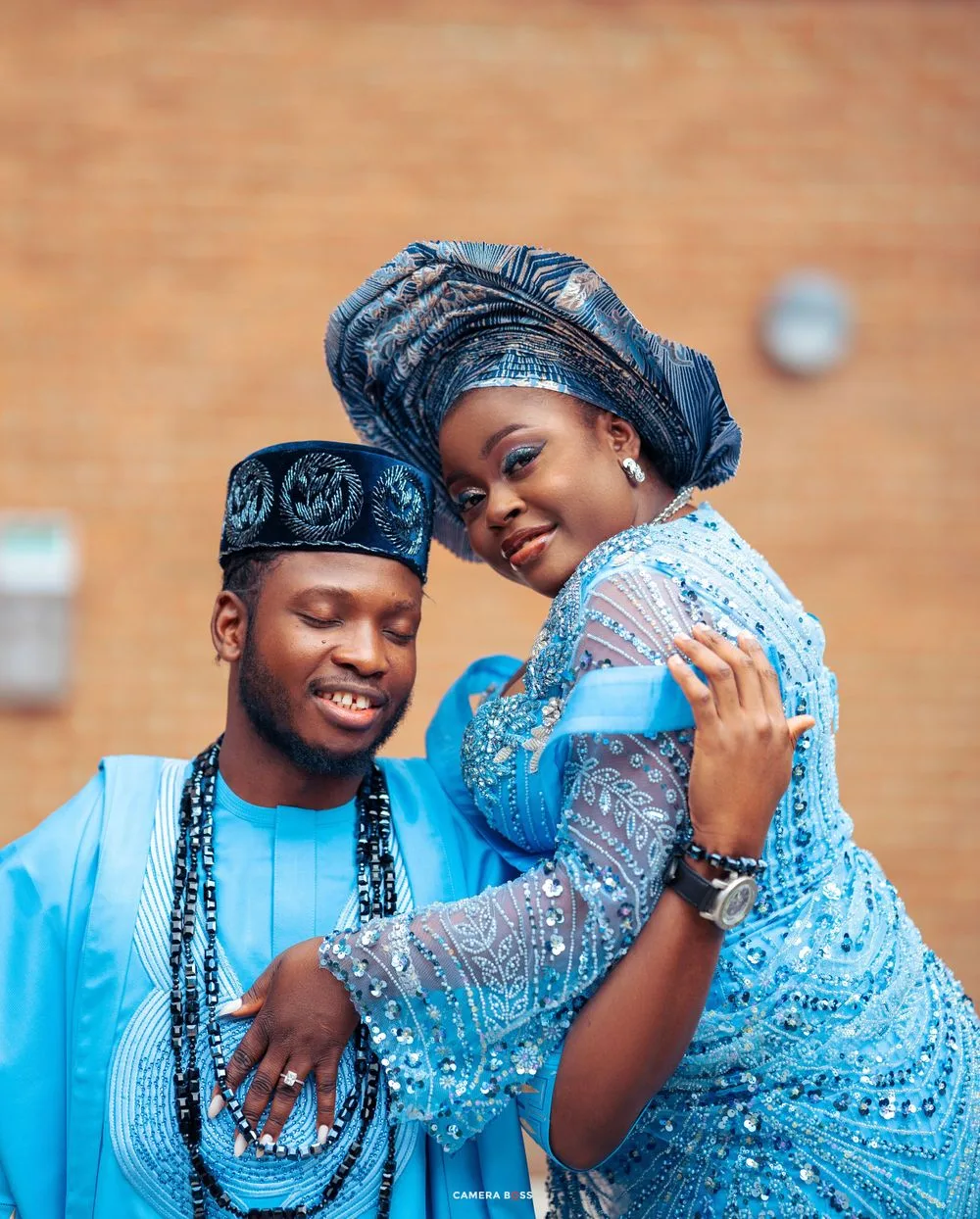 A couple in matching blue traditional Nigerian attire embrace and smile during a celebration.