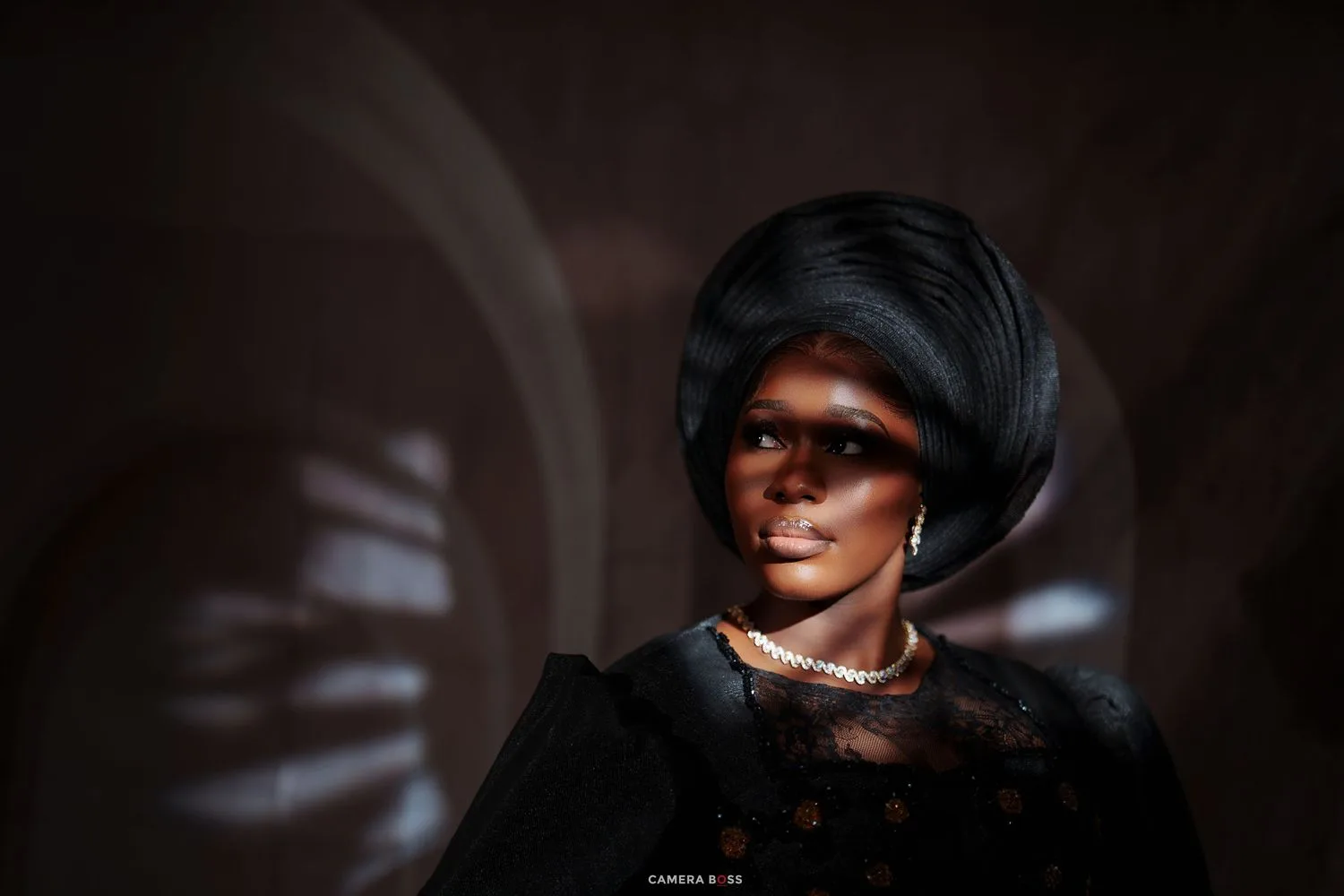Elegant woman in black lace dress with pearl necklace and sleek updo hairstyle, posed in dramatic studio lighting.