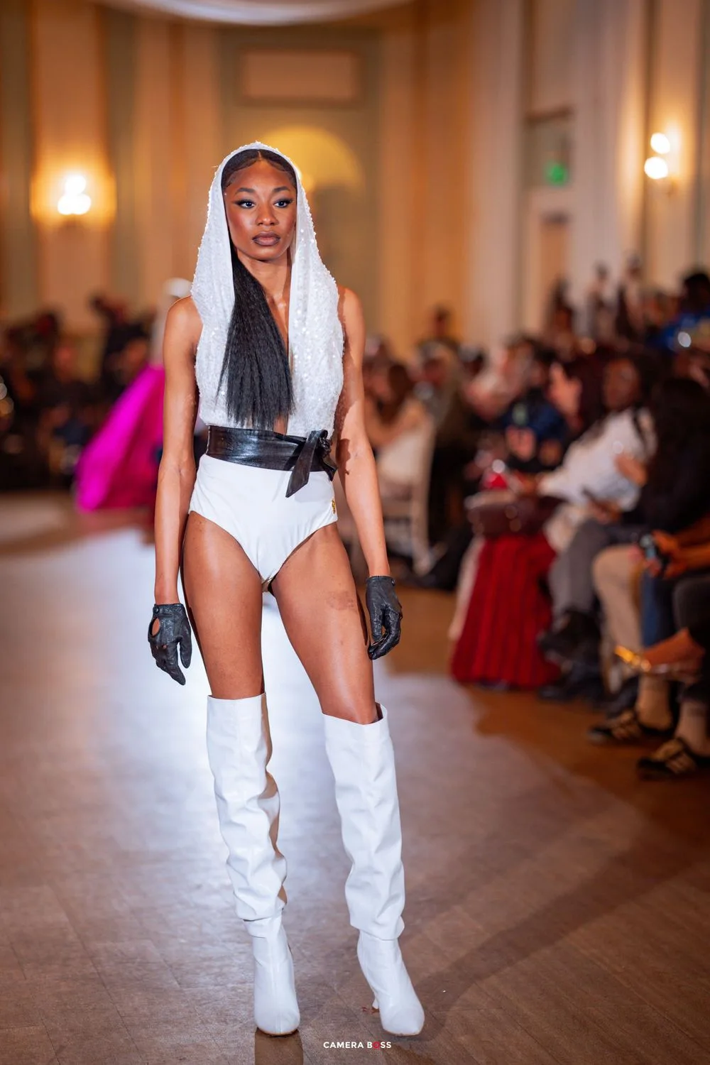 Model walks runway in white bodysuit with tall white boots and black belt during fashion show.