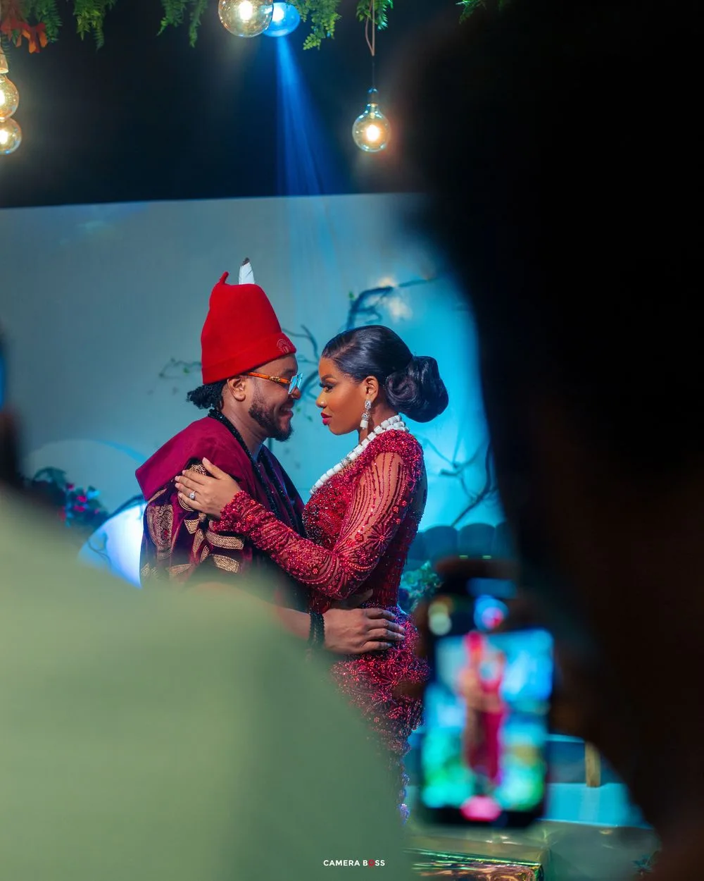Two people in festive attire share a romantic moment under dramatic blue lighting and hanging glass orbs.