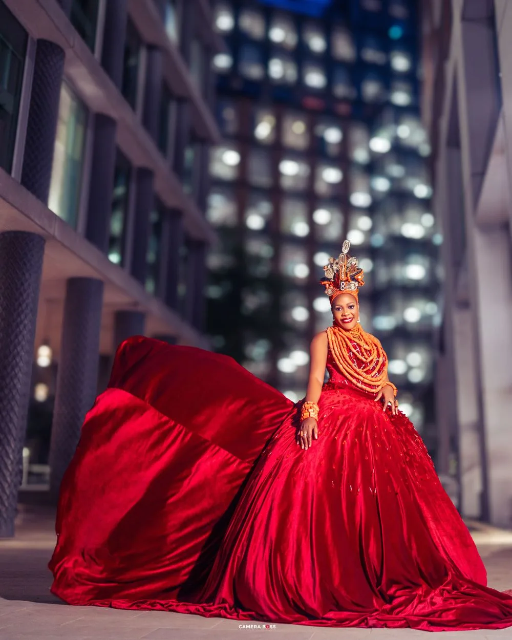 A figure in a dramatic flowing red gown and ornate headdress poses against an illuminated city building at night.