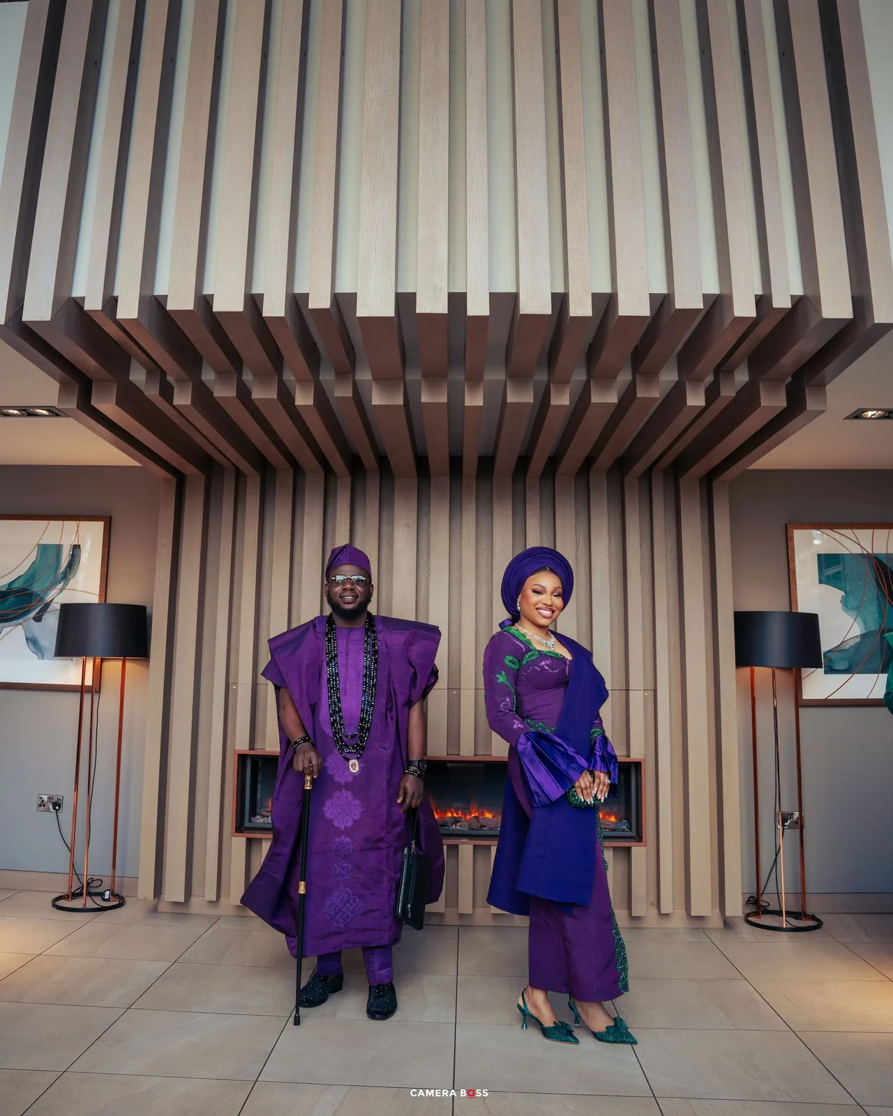 A couple dressed in coordinating purple and blue traditional African attire poses elegantly in a modern interior setting.
