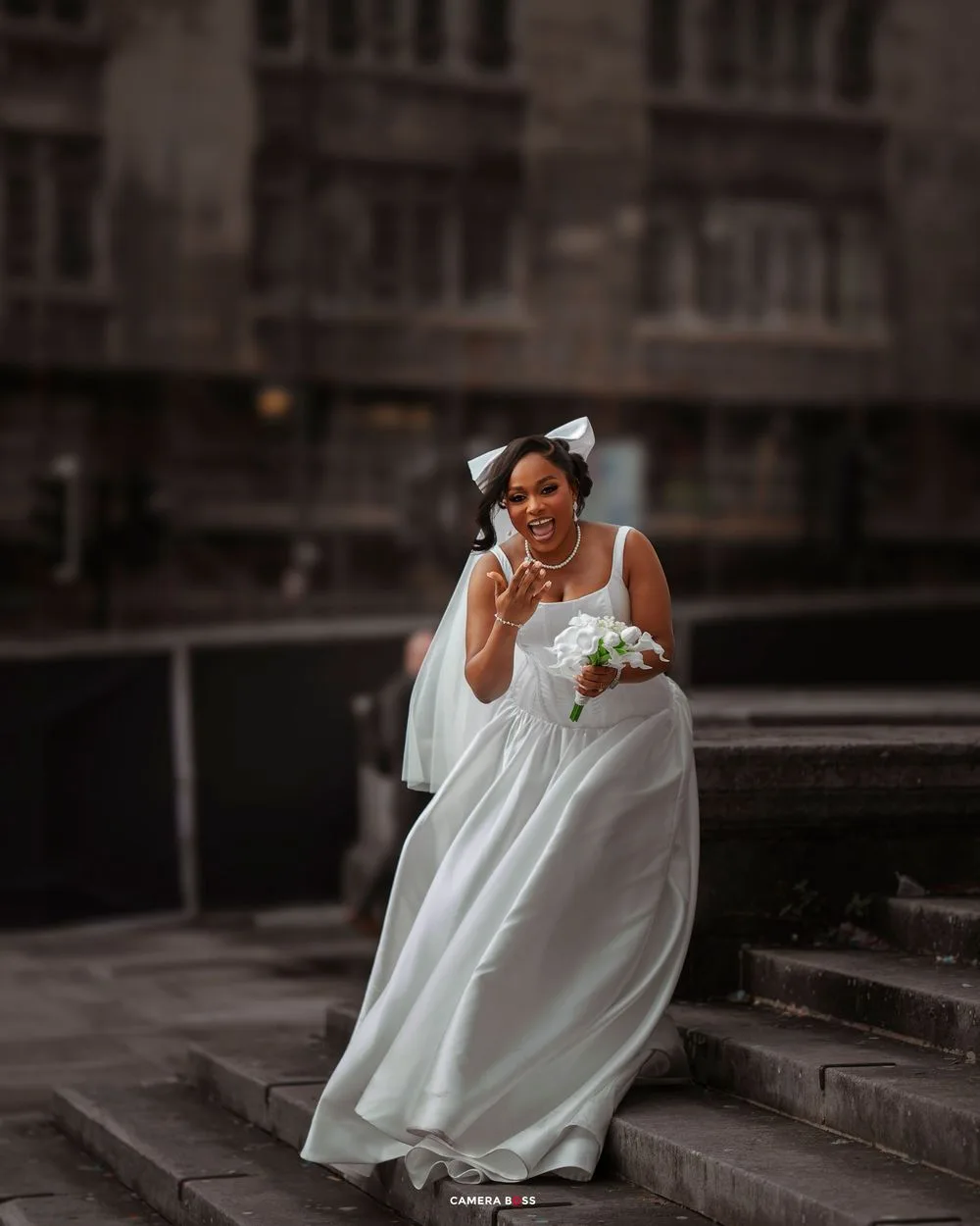A bride in a flowing white dress sits elegantly on concrete steps in a moody urban setting.