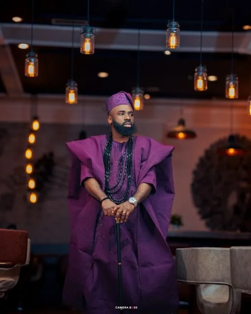 Traditional Nigerian agbada outfit in deep purple with matching cap, photographed under hanging pendant lights in a modern setting.