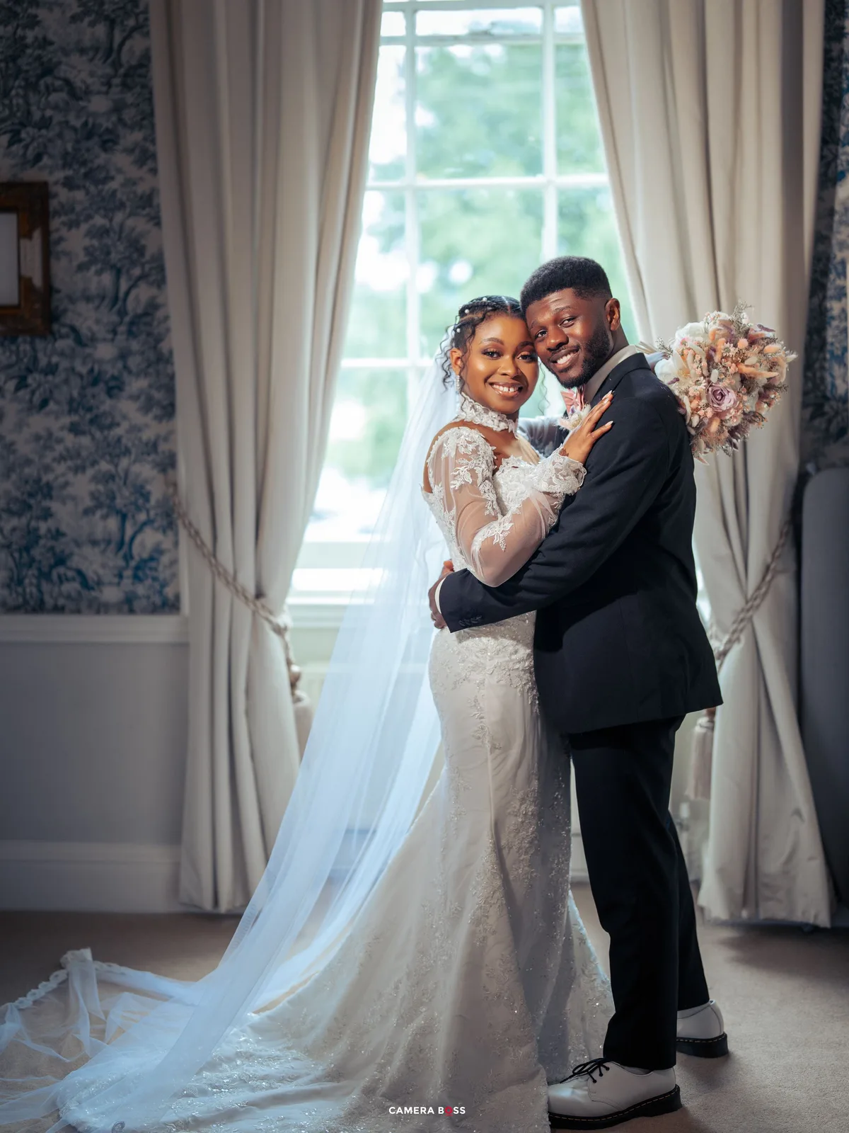A happy couple dances together in a elegant room with white curtains and flowing bridal gown.