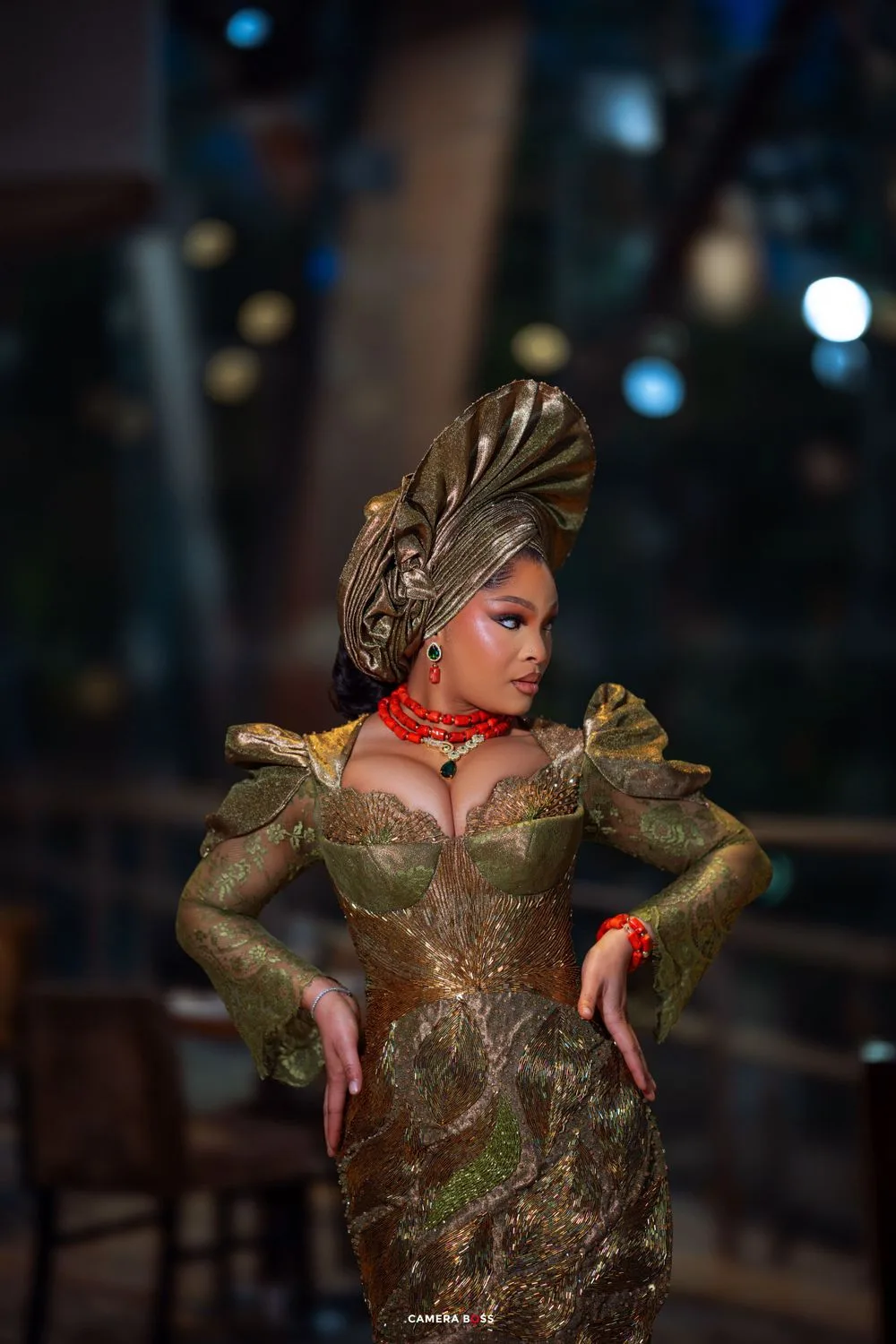 A figure in an ornate olive green dress with dramatic puff sleeves and an elaborate gold headpiece poses against a dark background.