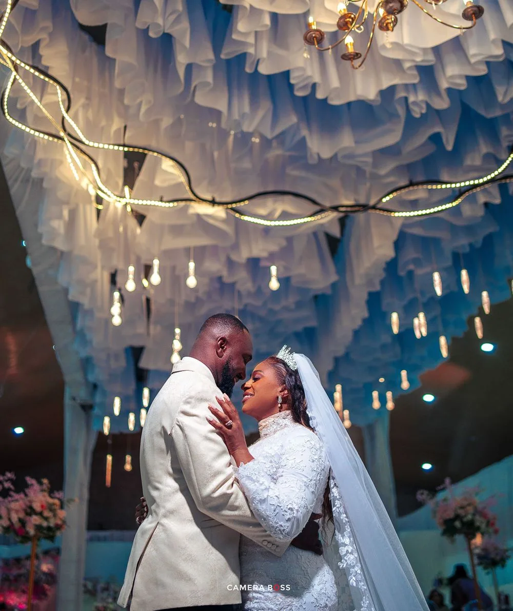 A romantic wedding moment captured under a dreamy ceiling adorned with hanging lights and draped white fabric decor.