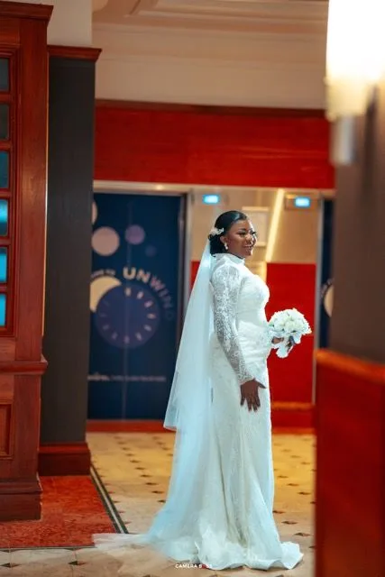 A bride in an elegant long-sleeved lace wedding gown and veil holds a white bouquet while posing in a red-walled interior.