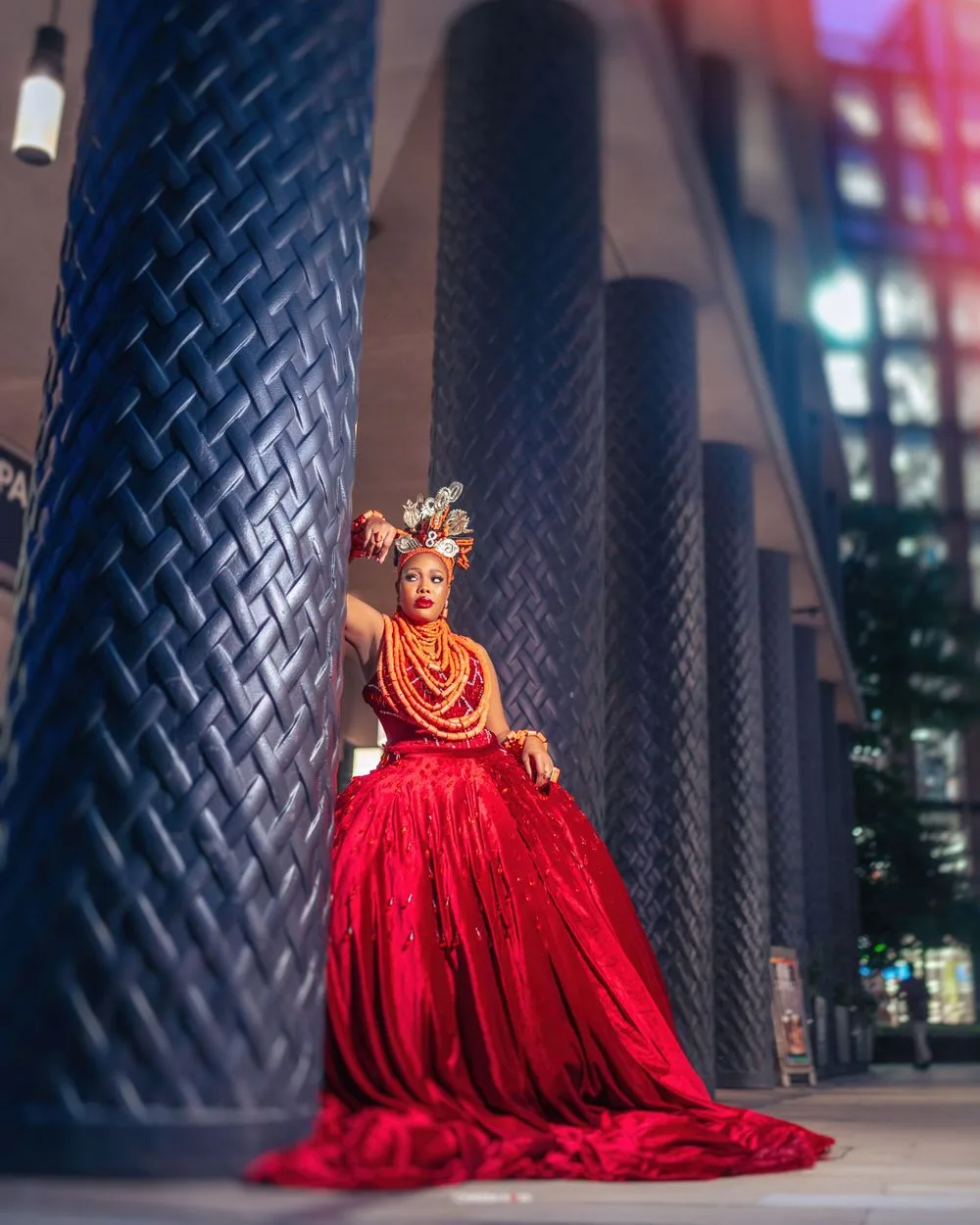 A stunning figure in a flowing red gown and ornate headpiece poses dramatically against tall textured black pillars.