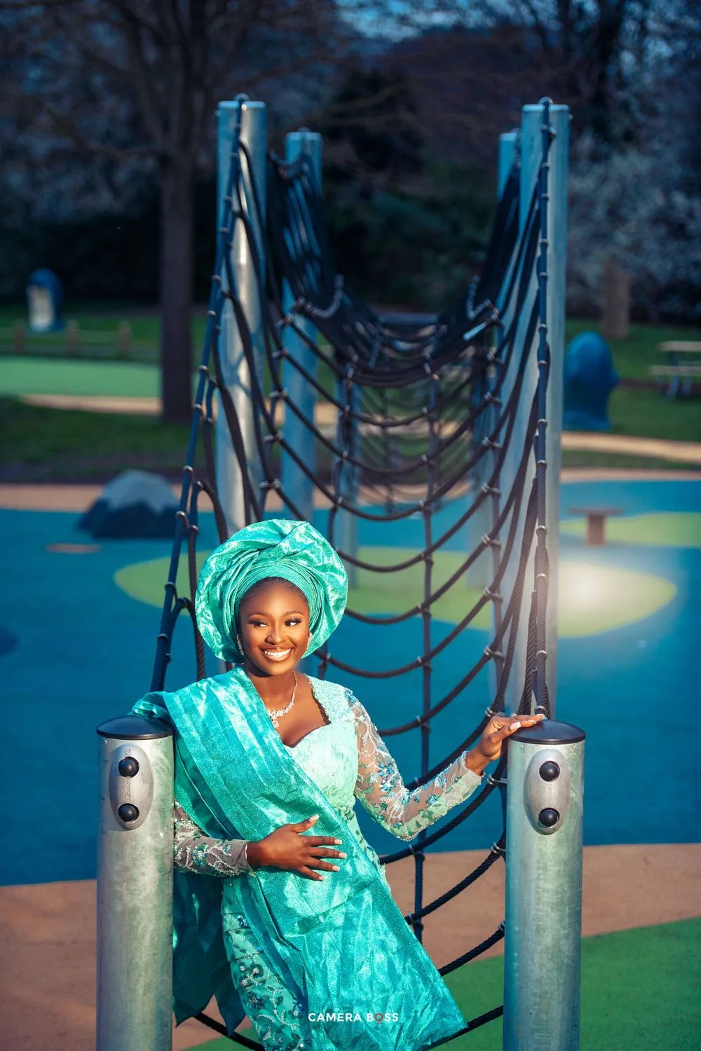 Person in vibrant turquoise traditional dress and headwrap poses by playground rope bridge at dusk.