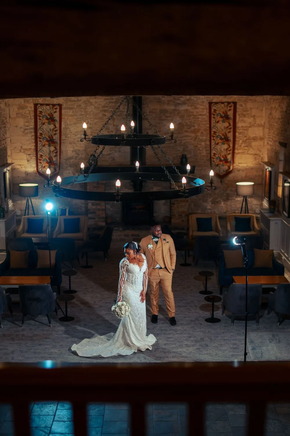 A couple stands in an elegant rustic venue with exposed brick walls, a wrought iron chandelier, and blue church pew seating.
