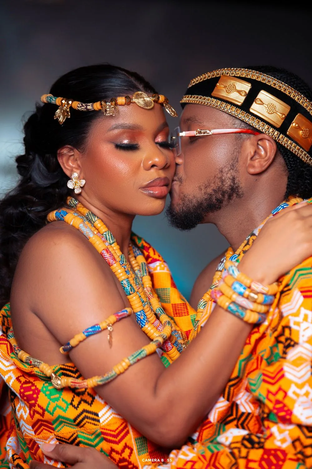 Two people in traditional African attire with colorful beads and kente cloth share an intimate moment in a studio setting.