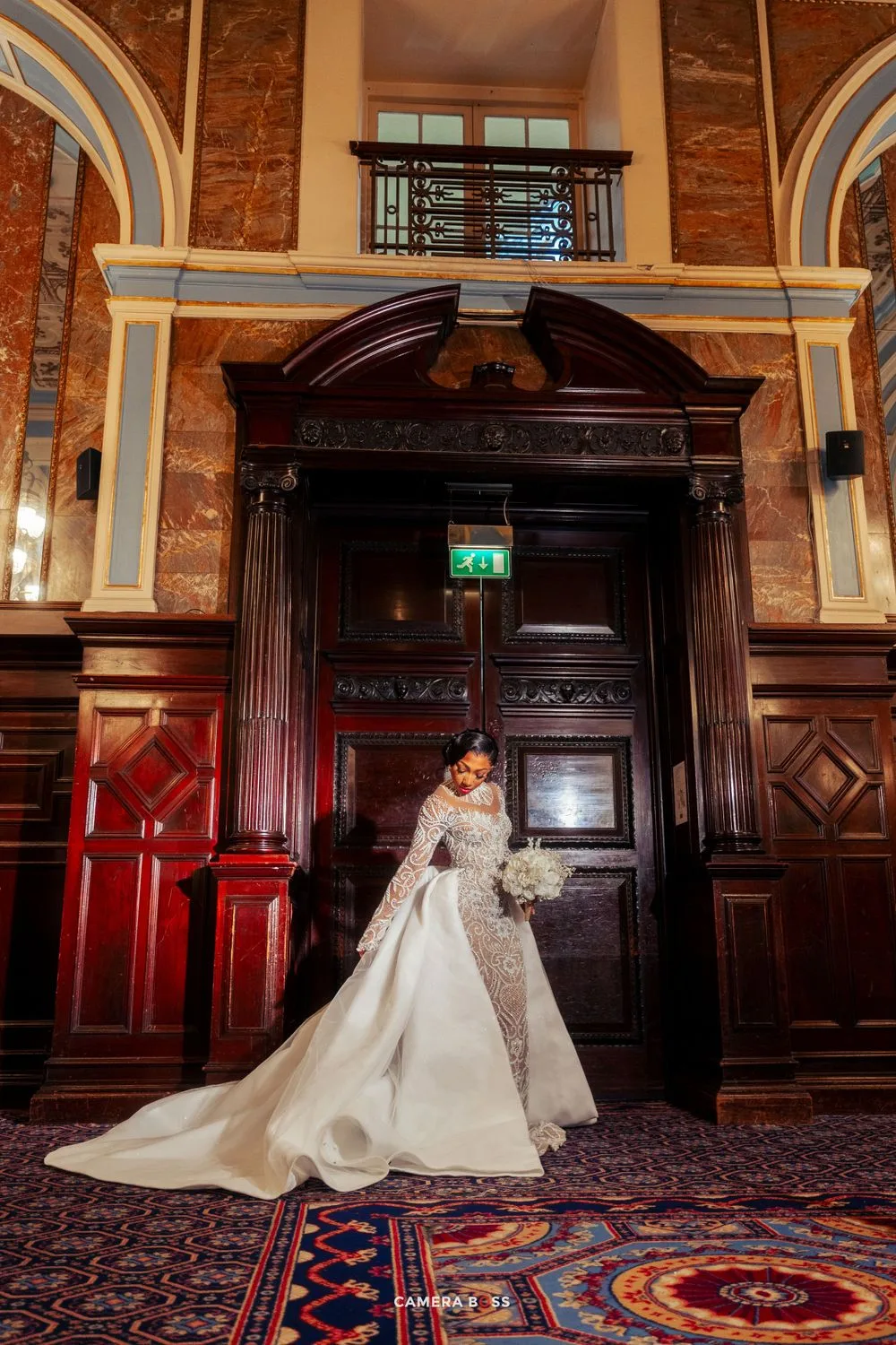 A bride in an elegant lace-sleeved wedding gown poses in front of ornate wooden doors inside a historic building with arches.