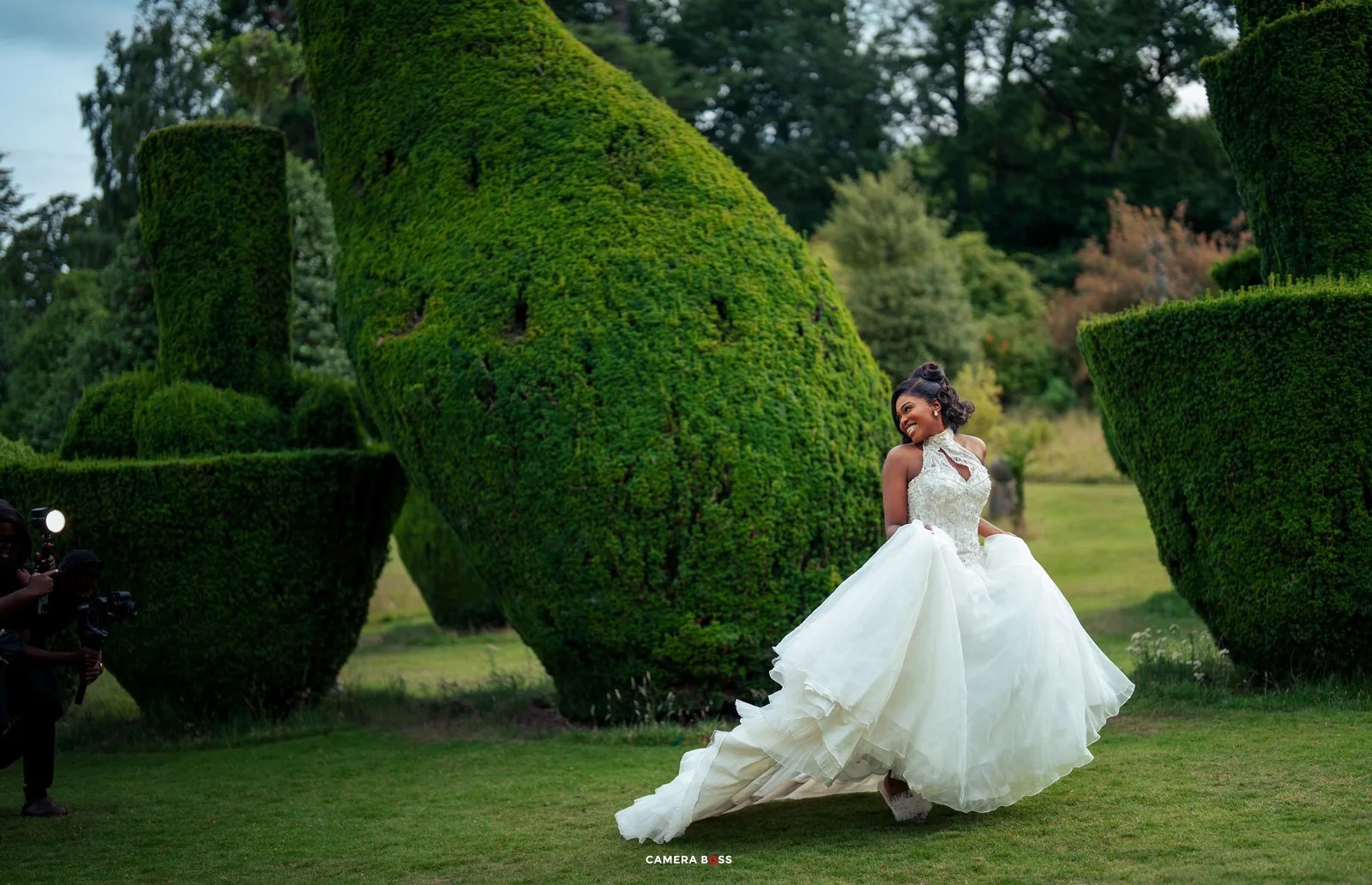 A couple in wedding attire poses in a garden with sculptured topiary hedges on a sunny afternoon.