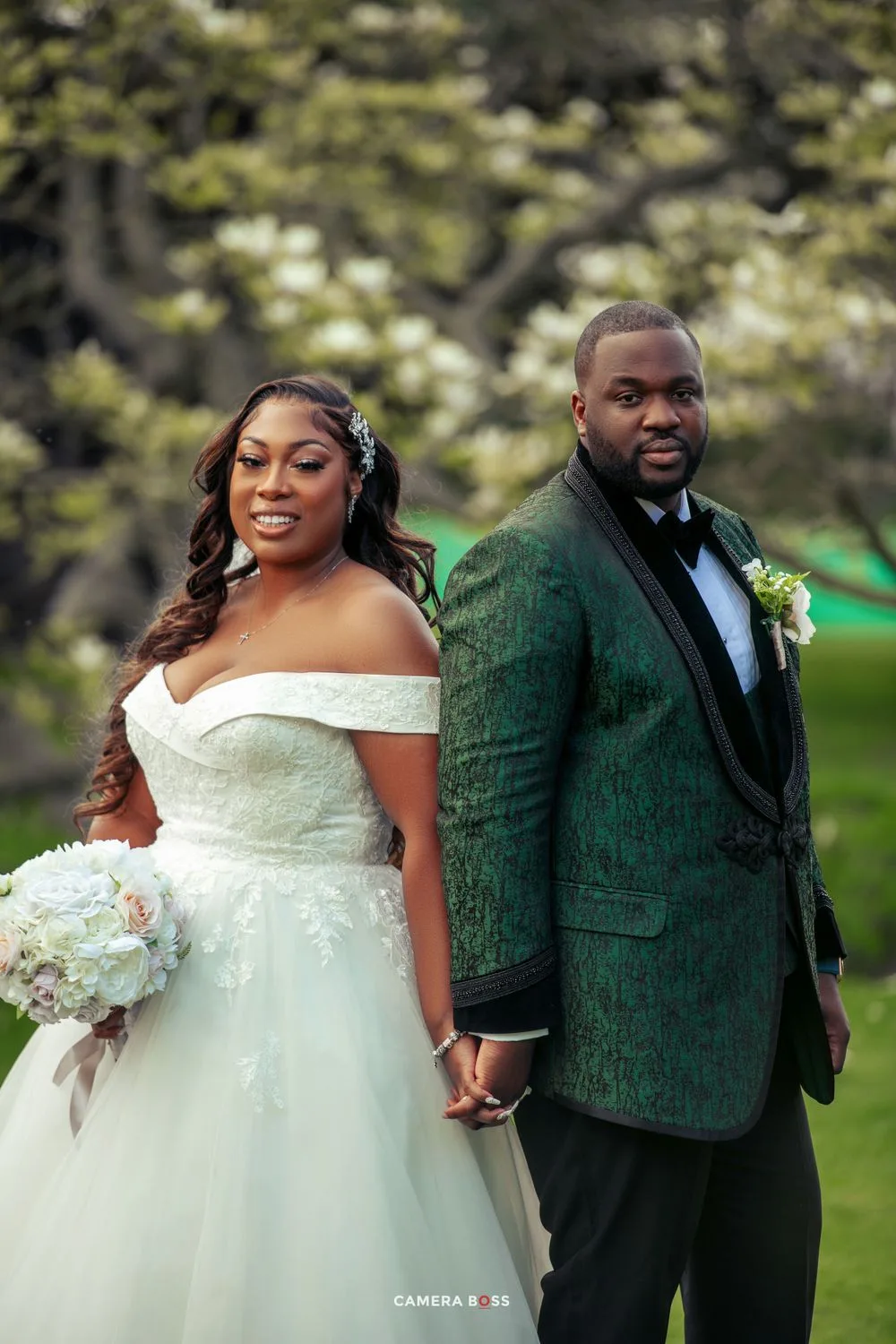 A couple in wedding attire pose together outdoors, with the bride in a white gown and groom wearing an emerald green jacket.
