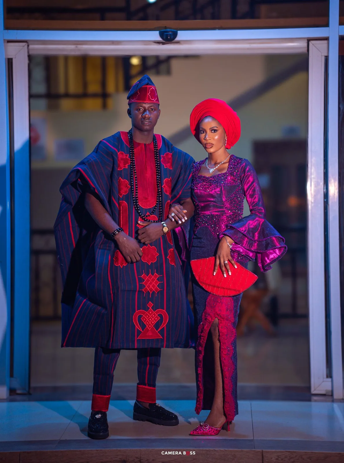 A couple in coordinated Nigerian traditional attire with navy blue and red colors pose elegantly in formal wear.