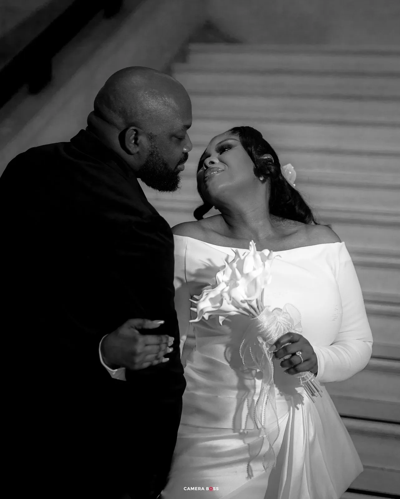 Black and white wedding photo of a couple in formal attire, bride holding bouquet, sharing a romantic moment on stairs.