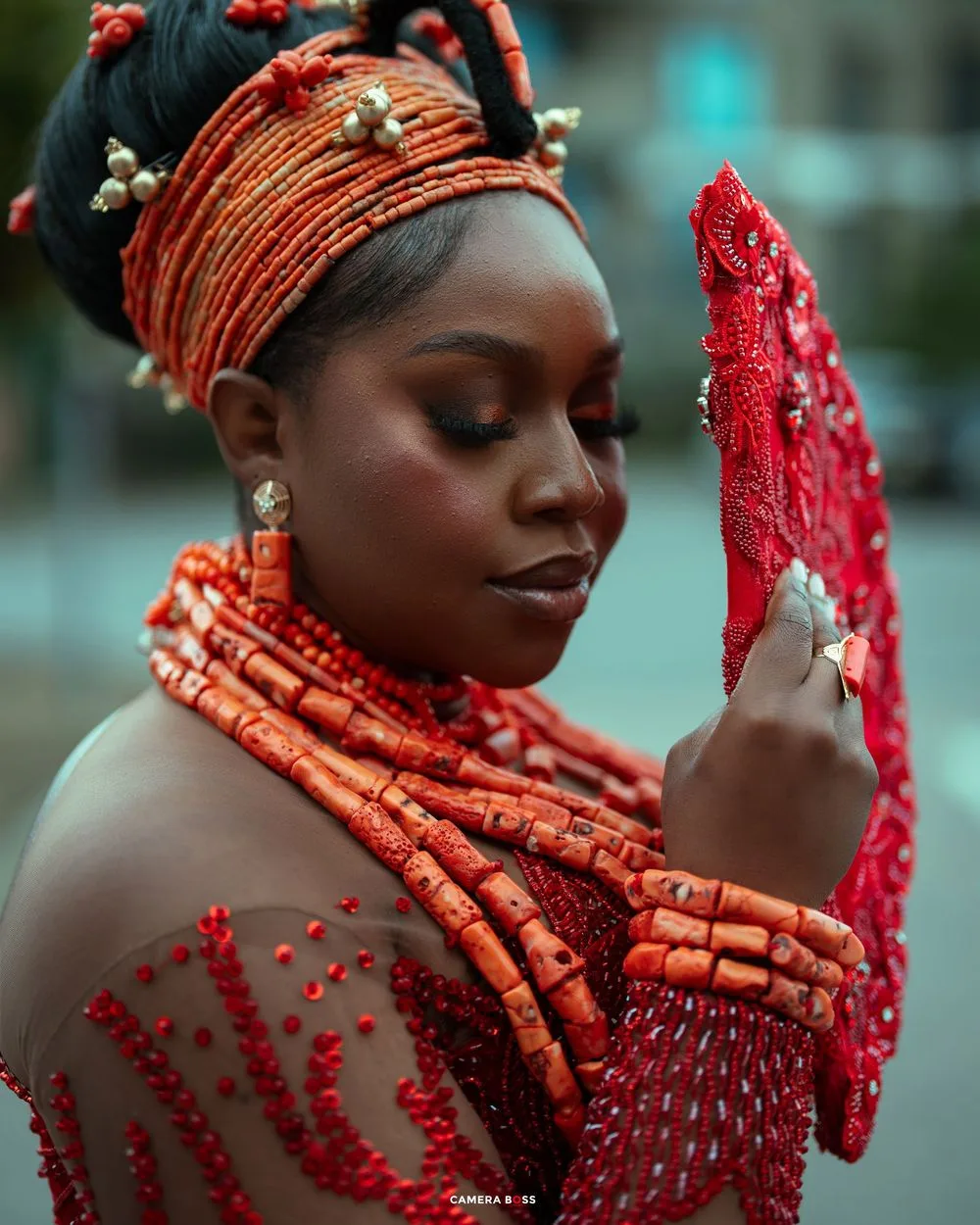 Close-up of woman in red Nigerian traditional coral beaded jewelry and headdress holding a red beaded fan.